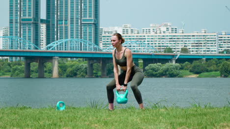 Fitte-Sportlerin-Beim-Kettlebell-Kreuzheben-Training-Im-Hangang-River-Park-Tagsüber,-Dongjak-Brücke-Im-Hintergrund-An-Einem-Sommertag-–-Vorderansicht