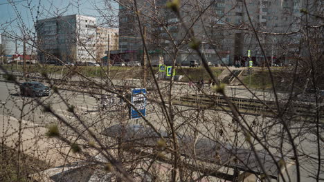 close view of a city street seen through budding branches, pedestrians are visible crossing the road, with buildings and road signs in the background