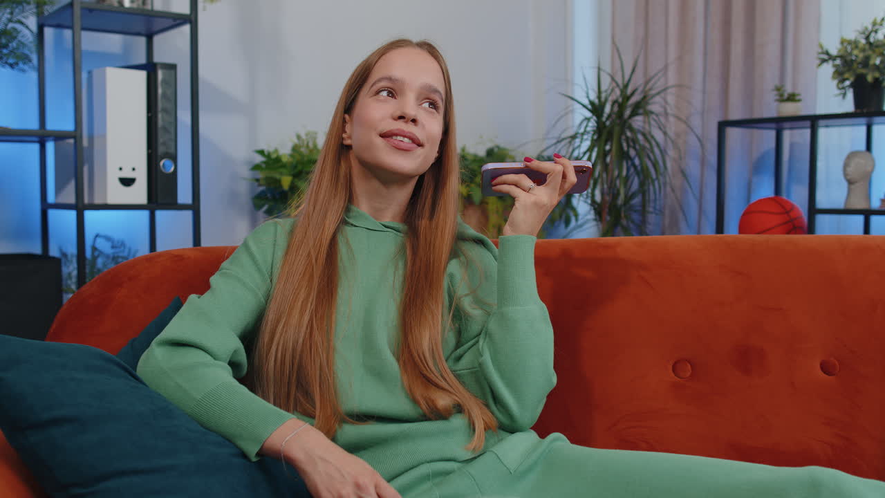 흥분 된 젊은 십대 소녀는 친구들과 전화 대화를 나누고 집에 있는 방에서 소파에 앉아 전화합니다.