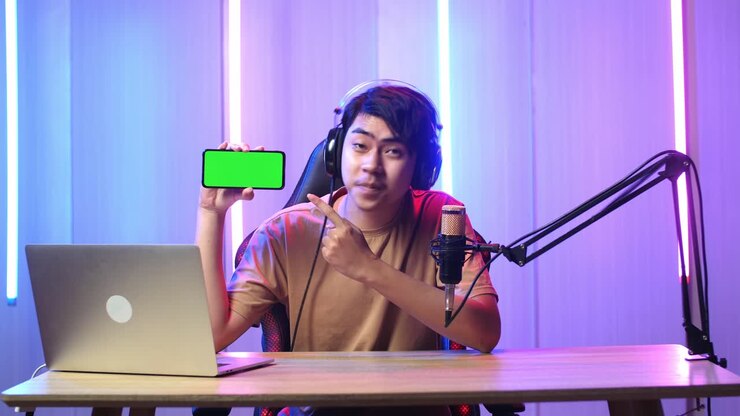 Uomo asiatico che tiene in mano un telefono cellulare con uno schermo verde di mockup mentre è in diretta streaming.