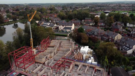 Establishing-aerial-view-crane-building-waterside-care-home-construction-framework-in-rural-British-village-next-to-fishing-lake