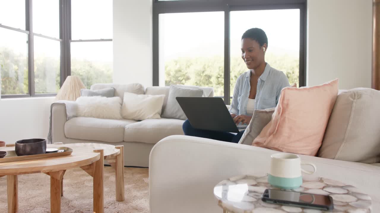 Happy african american woman sitting on sofa wearing earphones and using laptop, slow motion