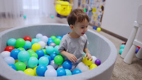 adorable dark-haired baby boy sitting in the dry basin. joyful kid throws the balls out of pool.