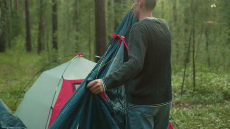 man in green knitted sweater holds tent cover while preparing to cover pitched tent in forest clearing, soft sunlight filters through trees as camping scene unfolds in natural woodland environment