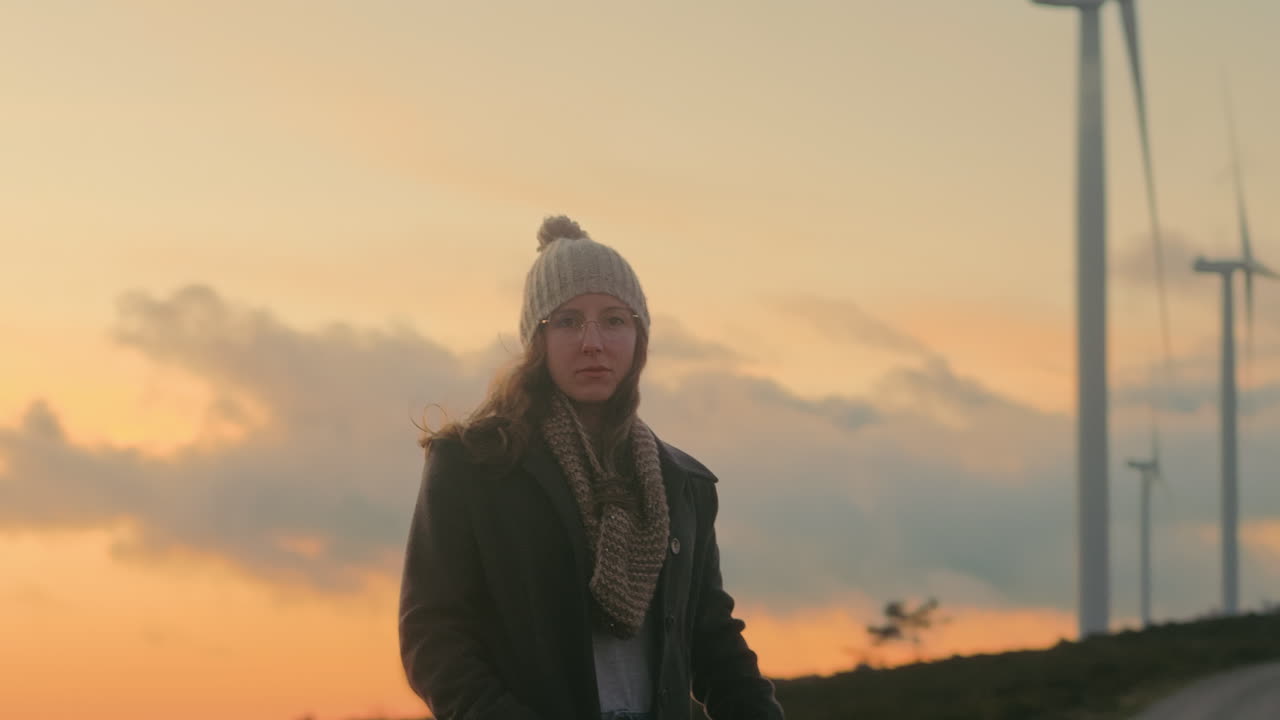 Blond kvinde med hue, briller, frakke og tørklæde står nær vindmøller i Portugal, nyder solnedgangen og den naturskønne skønhed. Symboliserer bæredygtighed, vedvarende energi, naturterapi. Filmisk optagelse.