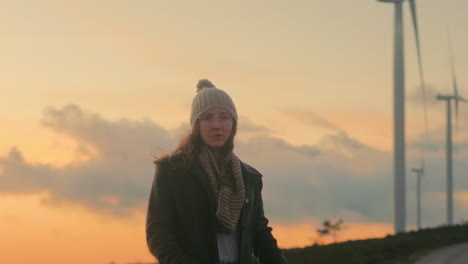 blonde woman in beanie, glasses, coat, and scarf stands near wind turbines in portugal, enjoying sunset and scenic beauty. symbolizing sustainability, renewable energy, nature therapy. cinematic shot.