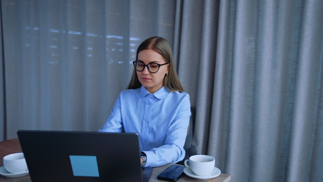 Beautiful young woman in glasses works on her laptop. Lady stands up, takes her cup and walks to the panoramic window in office.