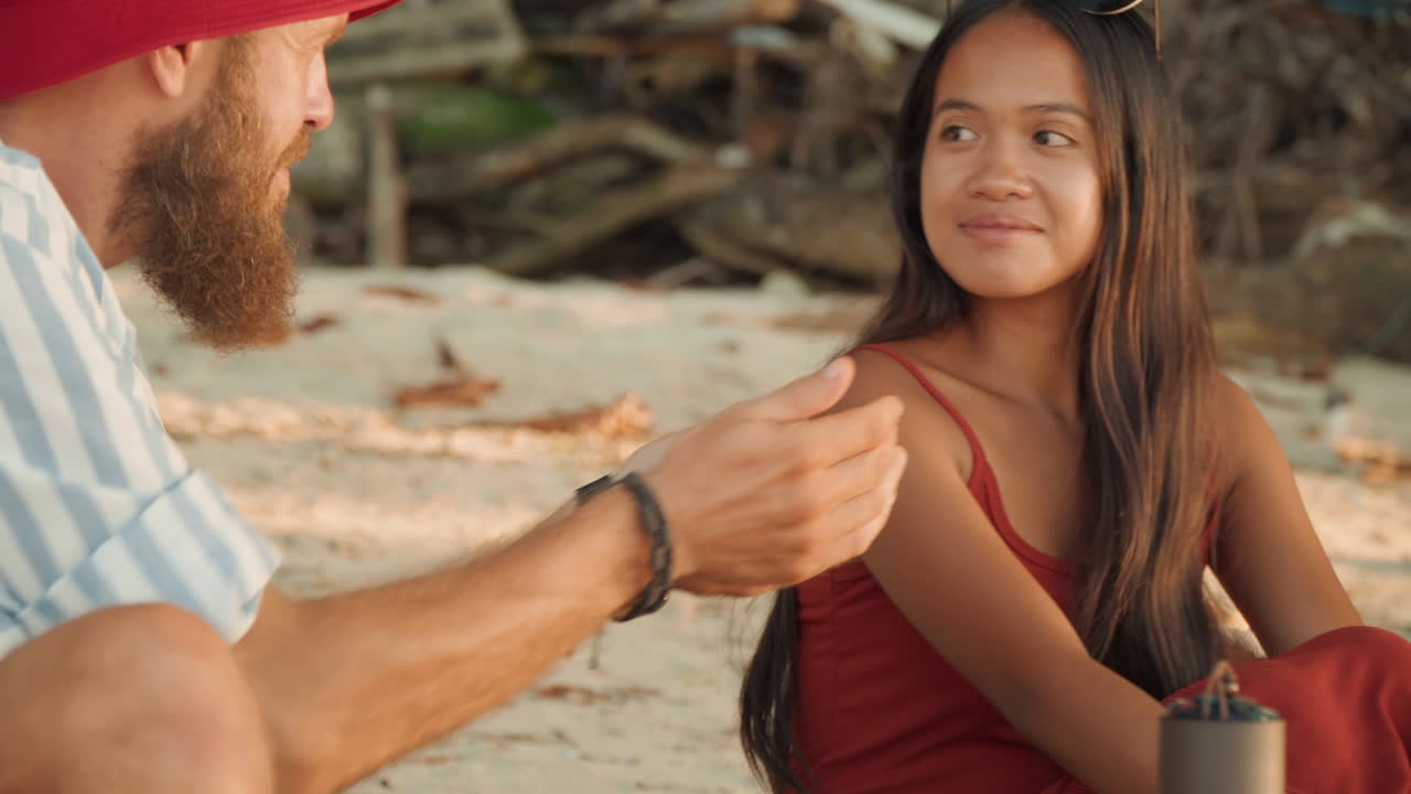 Intima cerimonia del tè in spiaggia con una coppia, donna in abito rosso e uomo seduti sulla sabbia che condividono tazze di ceramica su un vassoio rustico, bagliore dell'ora d'oro, conversazione calorosa, sfondo di legno trasportato dalla corrente, rituale rilassato a piedi nudi.