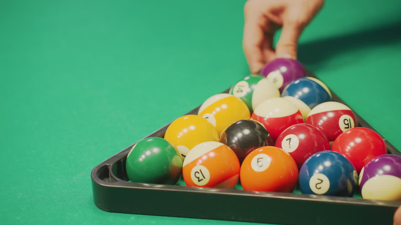 Close-up of hand rolling a well-arranged set of colorful billiard balls in triangle formation on green pool table. The polished balls reflect light, enhancing the precision and alignment