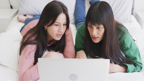 Happy-biracial-mother-and-adult-daughter-using-laptop-lying-on-bed,-slow-motion