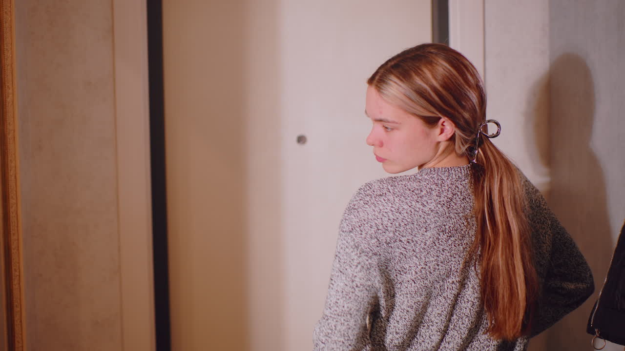 Young woman in gray sweater standing near doorway, thoughtfully looking at mirror reflection in hallway, adjusting sleeve with composed posture, showing elegance