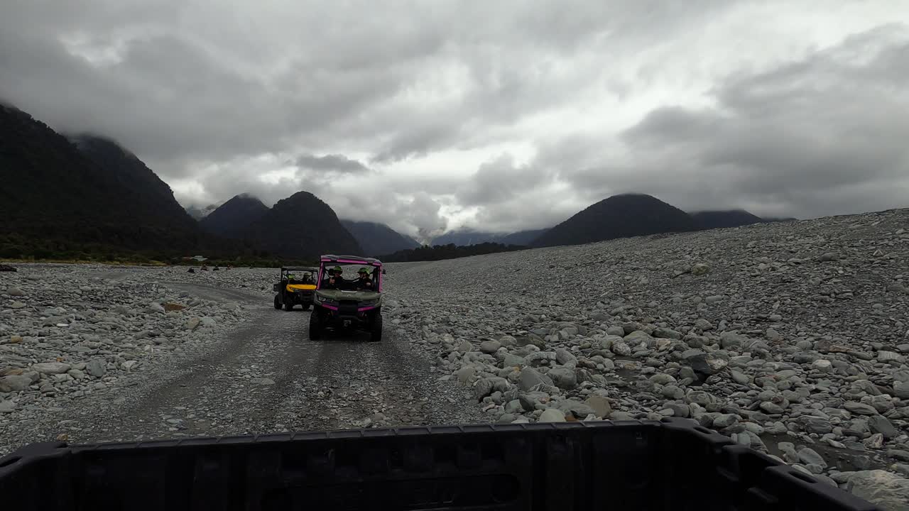 View from rear of car, Buggies and ATVs driving on rocky dirt road through dense rainforest under cloudy skies near Franz Josef, New Zealand.