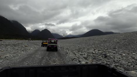 view from rear of car, buggies and atvs driving on rocky dirt road through dense rainforest under cloudy skies near franz josef, new zealand.