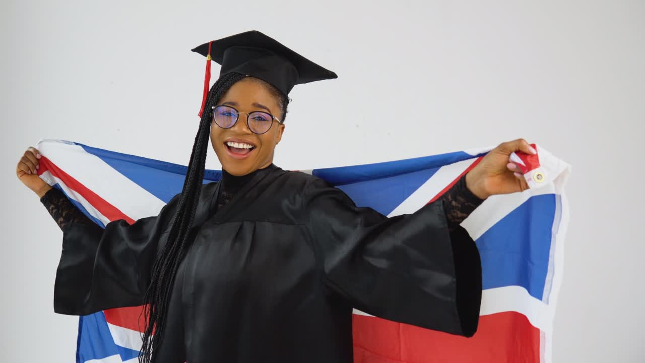 Happy stylish afro american woman student in graduate uniform is posing to the camera holding UK Flag on her shoulders