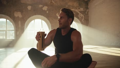 Close-up-shot-of-a-tired-male-athlete-in-black-sports-summer-uniform-drinking-water-from-a-special-sports-bottle-in-a-brick-sunny-hall