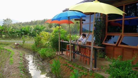 A-lone-female-sits-outside-at-a-small-rustic-local-café-with-colorful-umbrellas-in-a-remote-part-of-Nan-Province,-Thailand