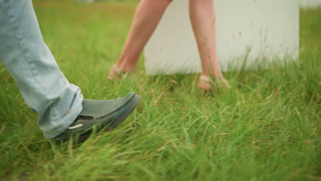 legs of two people walking through a green grass field. one is a man's leg wearing a black shoe, and the other is a woman's leg in sandals. focusing on the contrast between the footwear and the