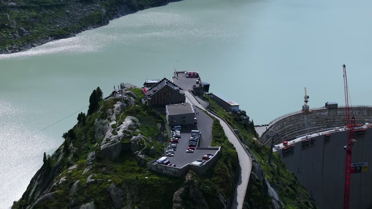 Aerial view showcasing the Grimselsee Lake Hotel and dam wall in Switzerland, with construction cranes visible amidst the mountainous landscape. Parallax Shot