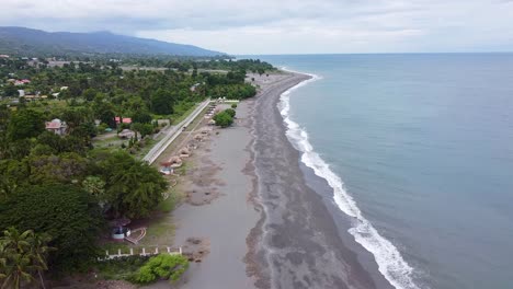 Aerial-drone-view-rising-over-black-sandy-beach-panoramic-landscape-with-waves-gently-rolling-onto-shoreline,-town-of-Liquica-near-capital-Dili,-Timor-Leste-in-Southeast-Asia