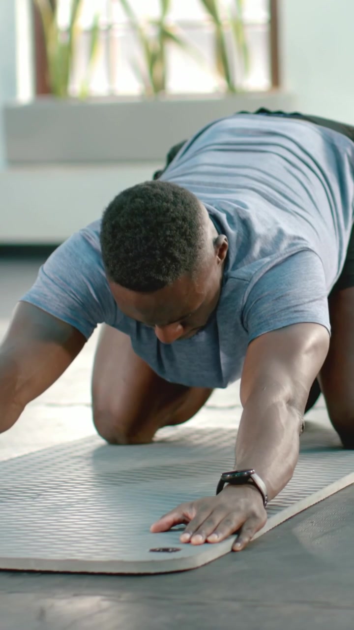 Vertical video: Kneeling man in blue top lowering chest to gray mat in gym, repeating for mobility