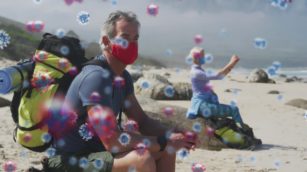 해변에서 백 ⁇ 과 함께 얼굴 마스크를 쓰고 있는 노인 부부 위에 떠다니는 코로나19 세포의 애니메이션