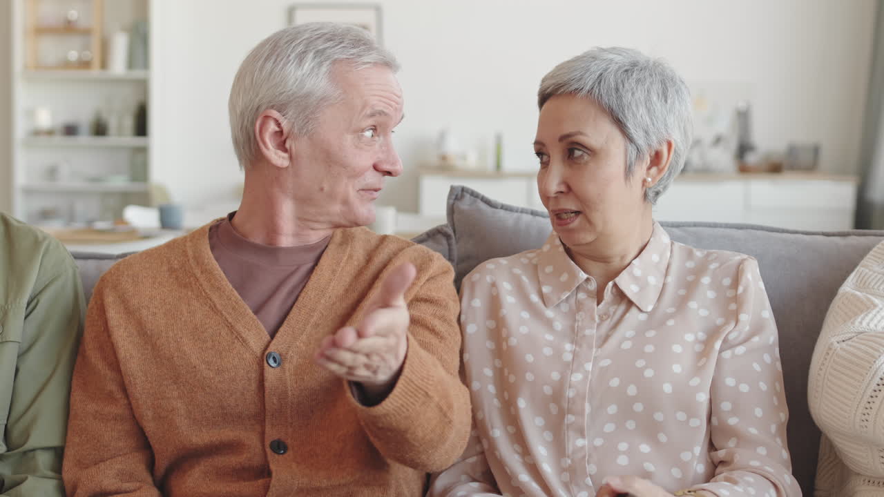 Waist up POV of older Caucasian man and Asian woman sitting on sofa at home, watching invisible TV, discussing it, smiling, pointing fingers