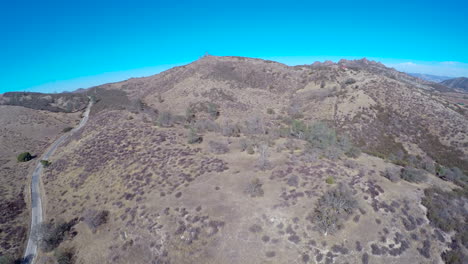 Una-Antena-Ascendente-Sobre-Un-Paisaje-Montañoso-árido-Y-Un-Automóvil-Que-Viaja-Por-Una-Carretera-Solitaria