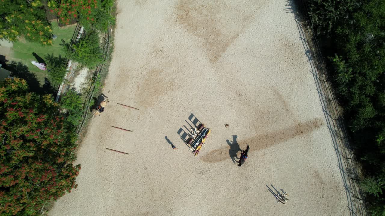 Ascending top down drone shot reveals a trainer gesturing to riders and the wide equestrian center area