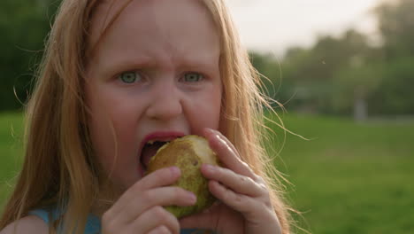 young girl using teeth to bite and cut pear in park, closeup portrait showing determined expression, messy juice on lips, sunlight soft background greenery and distant people enjoying outdoors