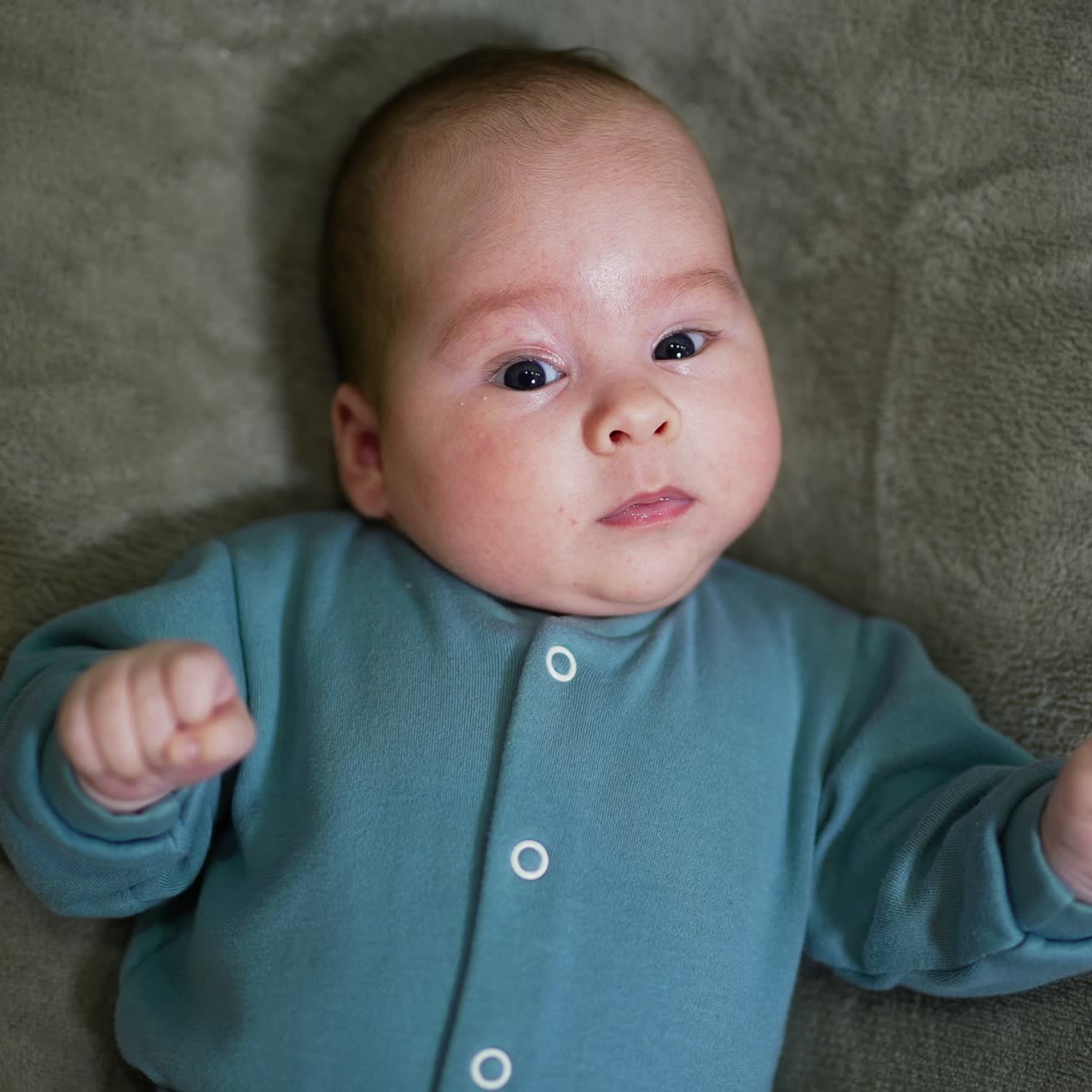 Bayi laki-laki mengenakan sweter biru berbaring dan melihat lurus ke depan. Anak kecil melihat kamera dengan terkejut dan berbaring diam. Tampilan atas. Close up