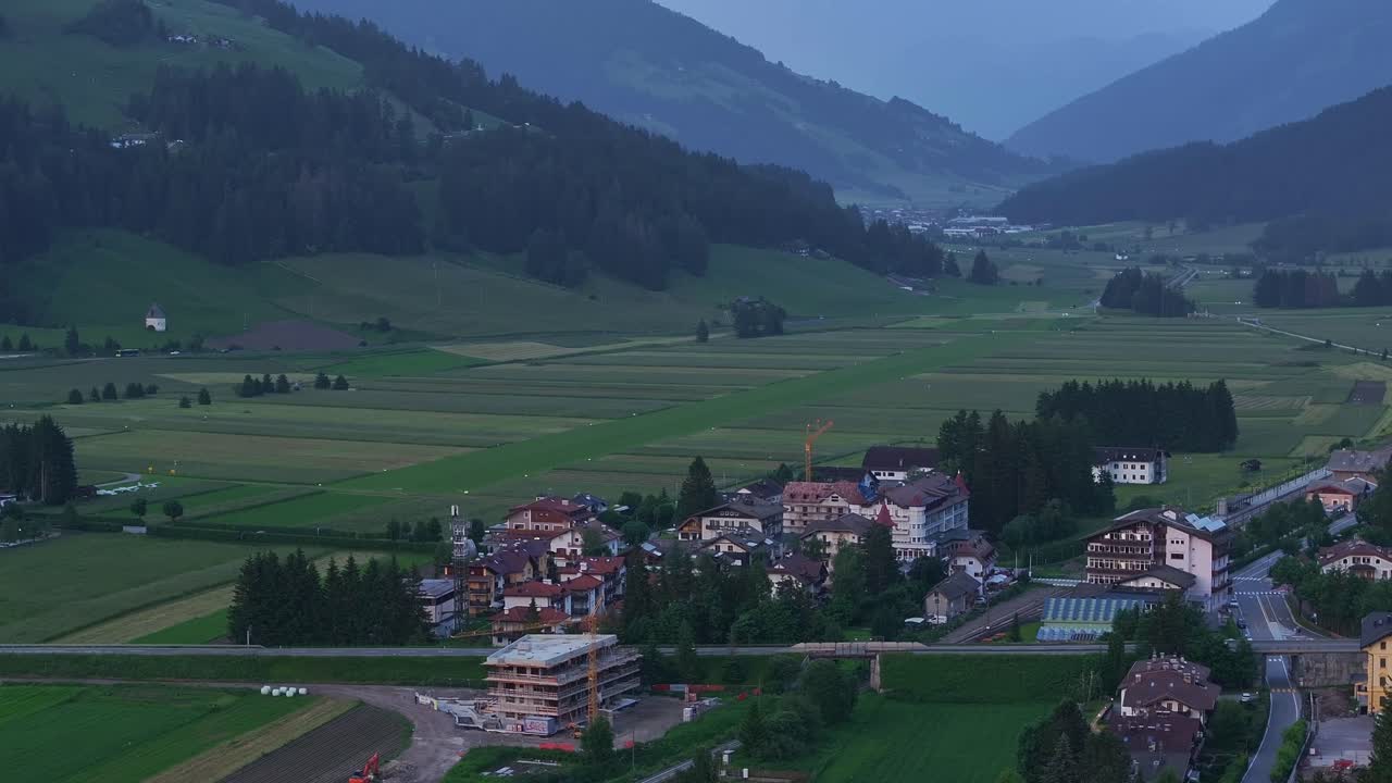 Wide aerial trucking pan above Toblach city apartment homes on edge of patchwork farmland at dusk