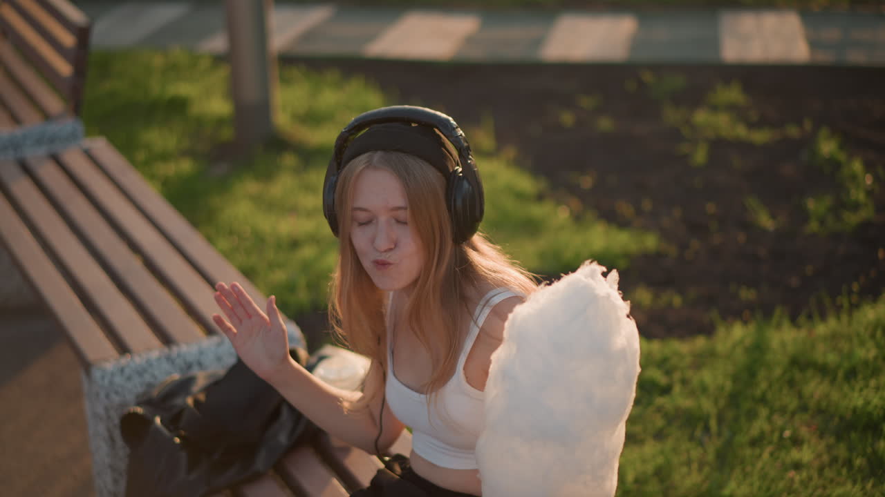 Junge, weiße Frau auf einer Parkbank, die mit Kopfhörern Musik hört, Zuckerwatte im goldenen Sonnenlicht genießt, entspanntes Lächeln, lässige Straßenkünstler-Ausstrahlung, urbane Picknick-Stimmung