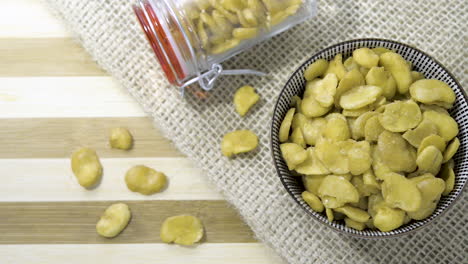 Slow-panning-shot-birds-eye-top-down-view-of-full-bowl-of-fava-beans-in-bowl-jar-across-wooden-table-with-woven-cloth