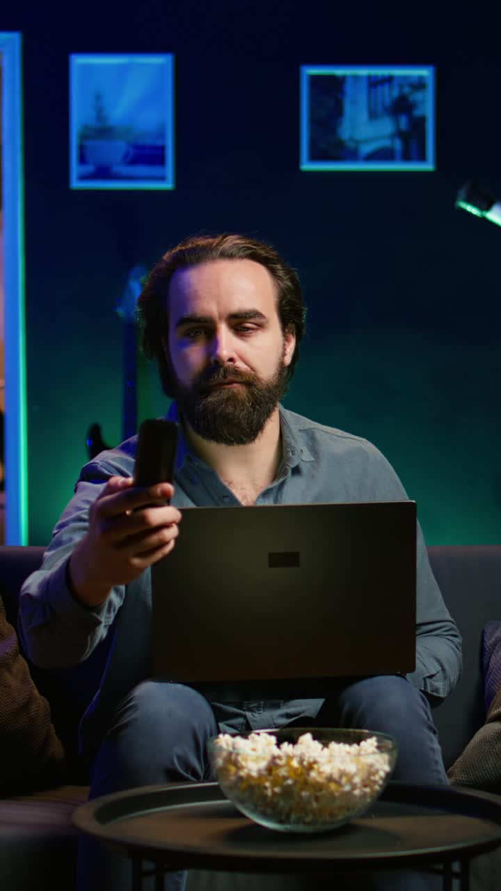 Vertical video Man typing on laptop, sitting on couch and watching TV