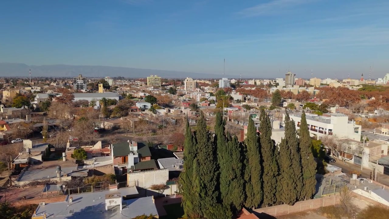 Panorama urbano di San Juan che si estende verso montagne lontane sotto un cielo azzurro limpido, Argentina, avvicinamento aereo che sorvola una barriera di alberi.