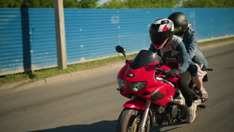 two relatives ride a red power bike, both wearing helmets, they are riding near place covered with blue zinc sheets with a building and parked car inside as they pass