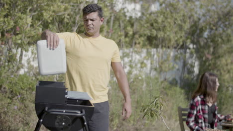 man standing at barbeque grill and waving lid to blow up fire