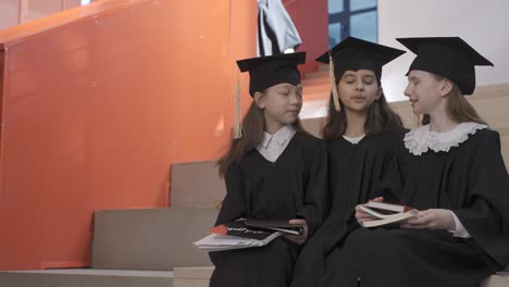 three happy little girls in cap and gown talking and holding their notebooks while sitting on stairs at the preschool graduation ceremony