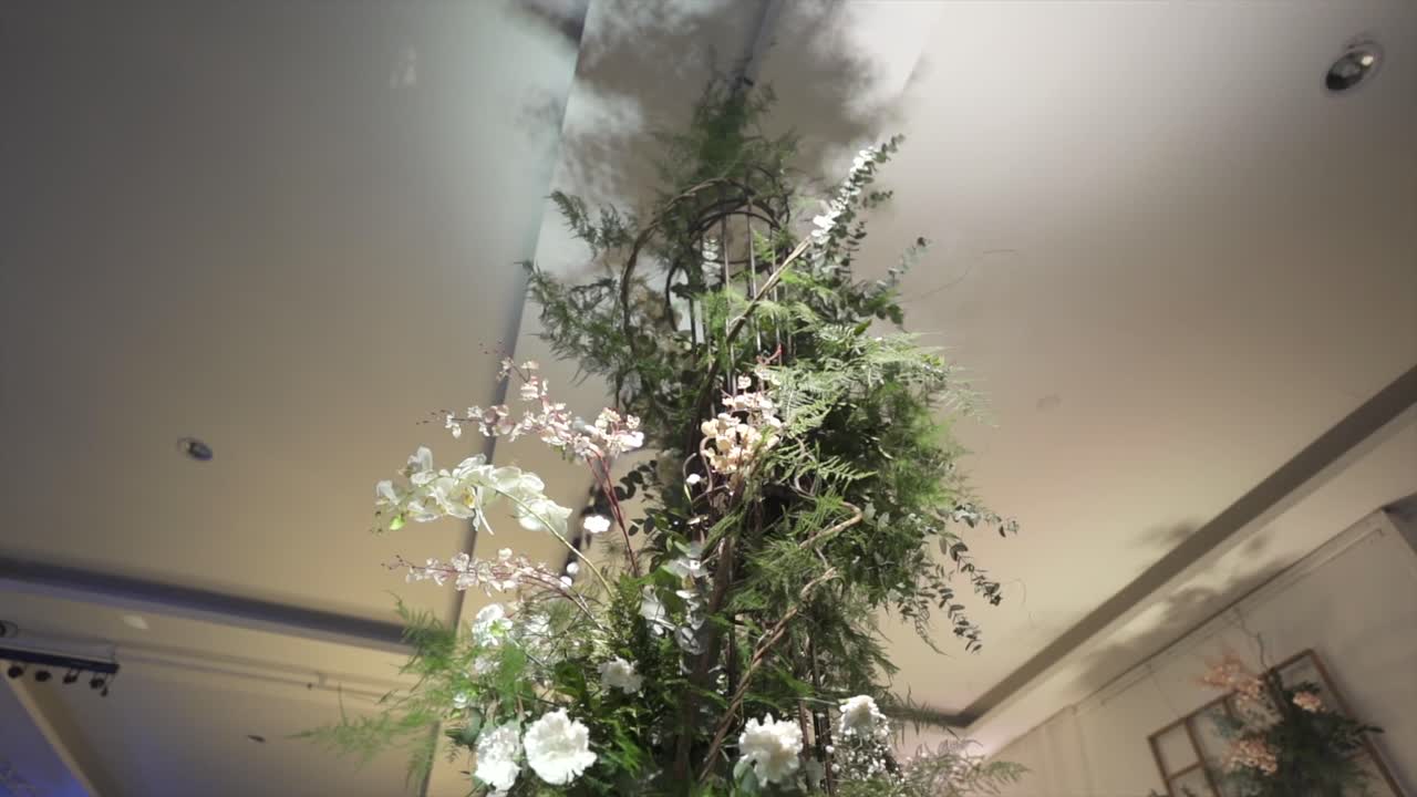 Tall floral arrangement of white roses, baby’s breath, and greenery rises toward the ceiling, captured in ascending shot that highlights its elegant structure and soft light in formal indoor setting