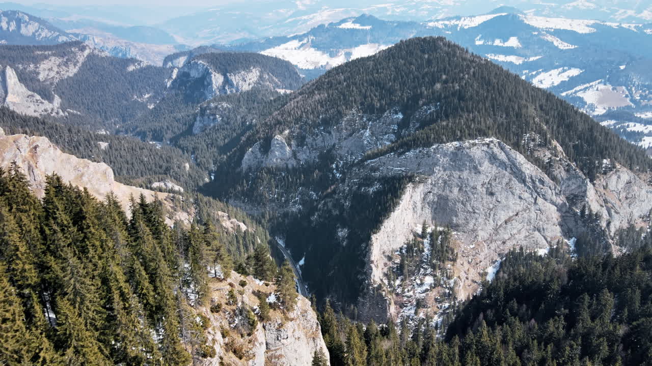 Luftaufnahme mit einer Drohne der Karpaten im Frühling, Rumänien. Felsige Hänge und ein Tal mit Straße, der Wald ist teilweise mit Schnee bedeckt