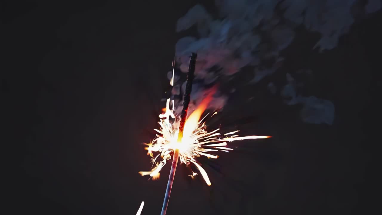 Close-up video of a lit sparkler against a dark background, capturing bright sparks and smoke