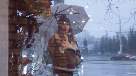 young girl holding umbrella walking past cafe, looking through glass window on cold overcast day, wearing black knit cap, brown shearling jacket, passing cars, and wet street in background