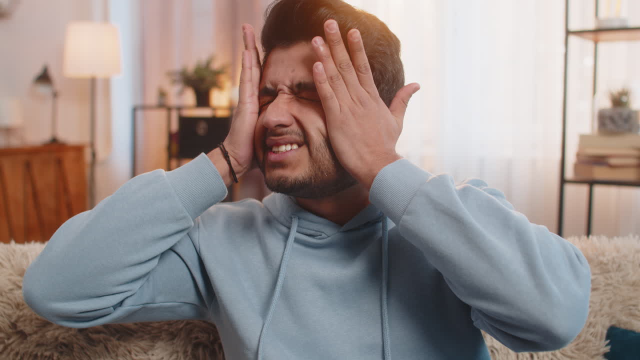 Indian young man on sofa rubbing temples to cure headache suffering from stress and migraine at home