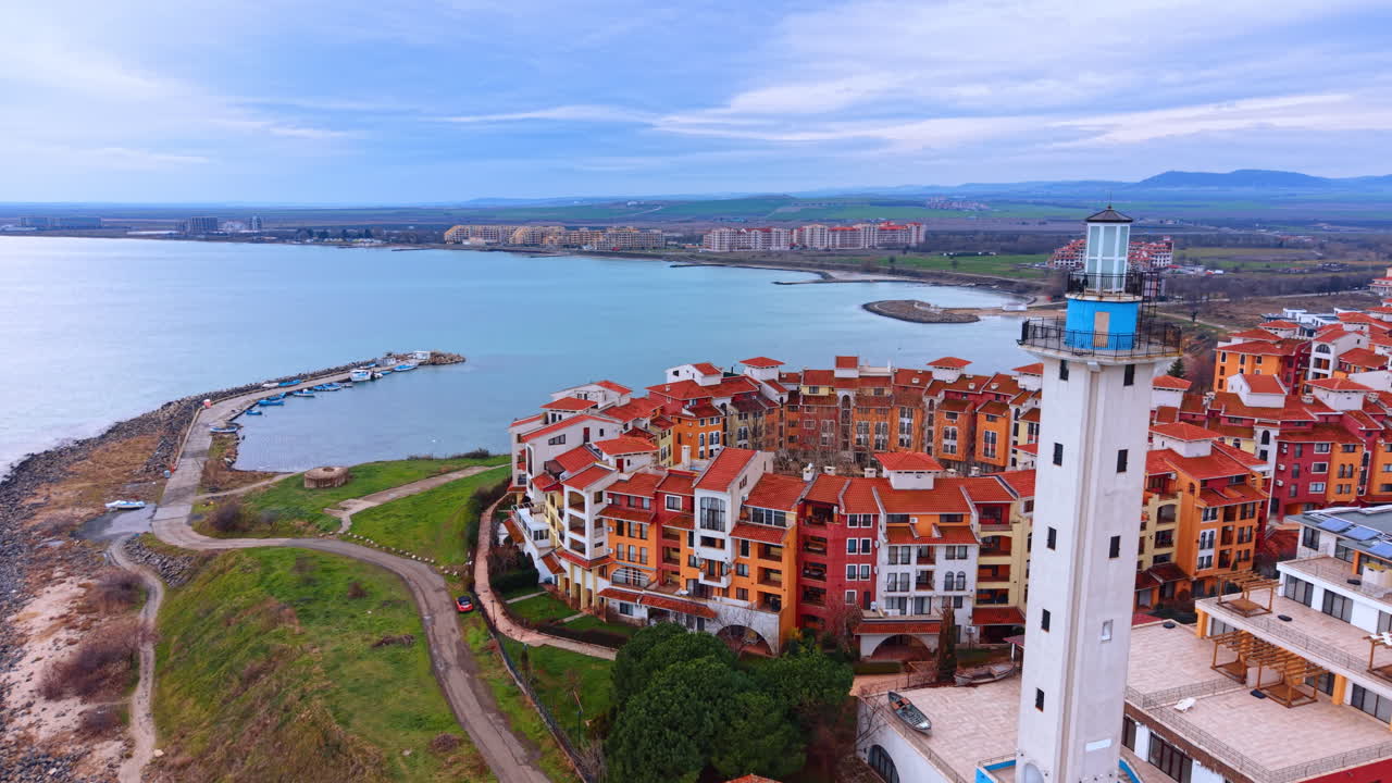 Vista aerea di un faro bianco e di edifici di un resort costiero. Ripresa con drone di una torre del faro accanto a un complesso di appartamenti con tetto arancione in riva al mare.
