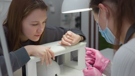 client and nail technician examine red nail polish bottle under salon light, checking if color matches previously used shade. technician wears pink gloves and mask, ensuring hygiene and precision