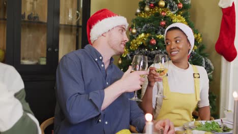 Fröhliche-Gruppe-Verschiedener-Freunde-In-Weihnachtsmützen,-Die-Das-Essen-Feiern-Und-Zur-Weihnachtszeit-Mit-Wein-Anstoßen