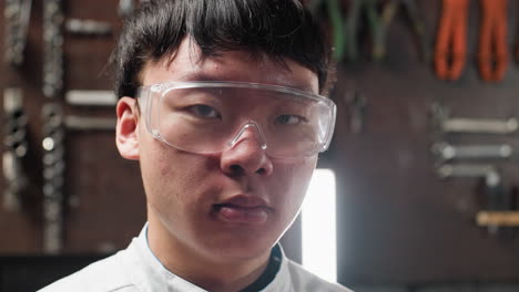 lab scientist wearing protective goggles blinks eyes with a thoughtful expression in mechanical workshop, background features industrial tools, bright lighting, and machinery