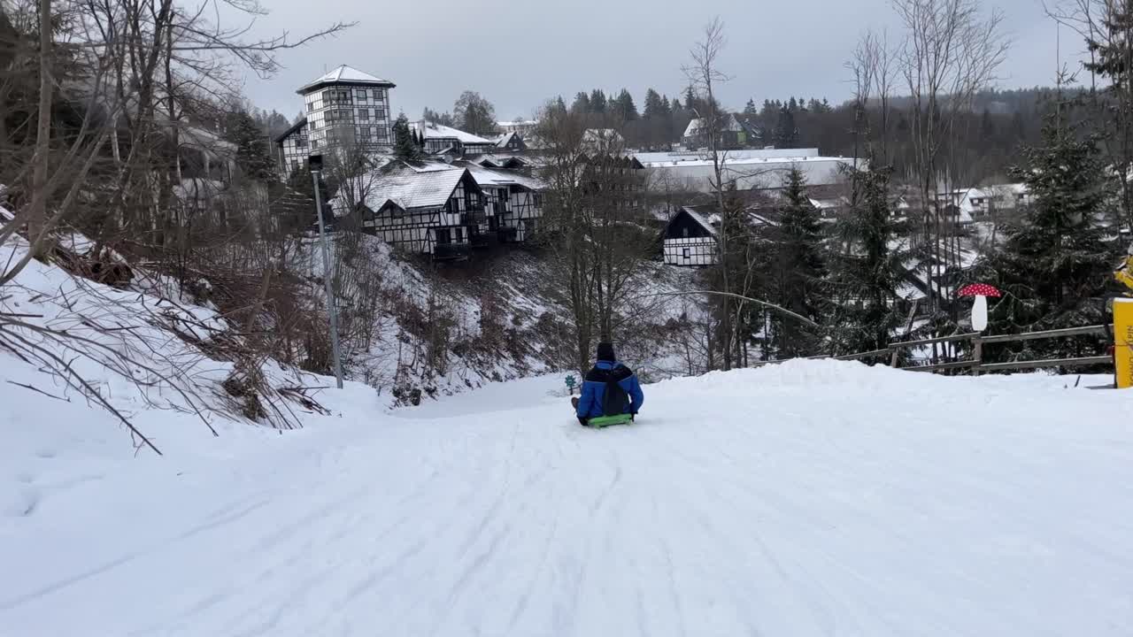 ciclista cuesta abajo con una chaqueta azul y negra cabalga en un trineo por una colina cubierta de nieve con pequeñas casas en el fondo y un bosque invernal en el borde