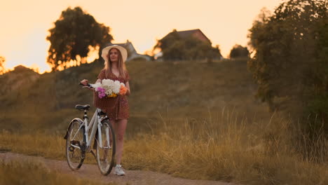 Young-beautiful-blonde-girl-in-summer-in-dress-and-hat-walking-on-road-with-bike-and-flowers-in-slow-motion.