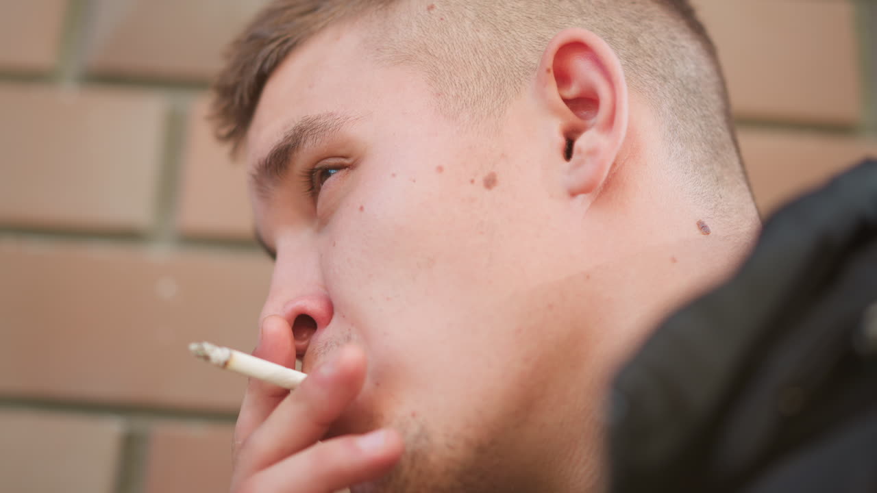 close up of man puffing smoke from cigarette while leaning against wall, face tilted with calm focused gaze, soft light revealing skin texture and moment of quiet reflection